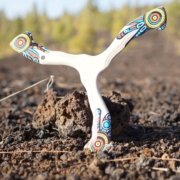 Spalvingas bumerangas „Šamanas“ padėtas ant tamsių vulkaninių akmenų gamtoje.