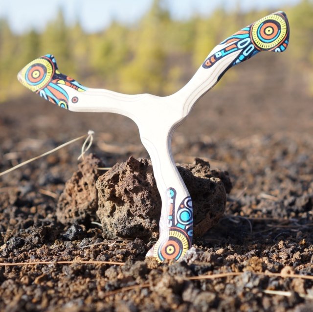 Spalvingas bumerangas „Šamanas“ padėtas ant tamsių vulkaninių akmenų gamtoje.