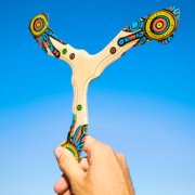 Vyro ranka laiko spalvotą bumerangą „Šamanas“ iškeltą prieš giedrą mėlyną dangų.