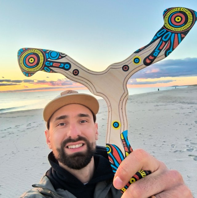 Besišypsantis vyras su kepure laiko bumerangą „Šamanas“ paplūdimyje.