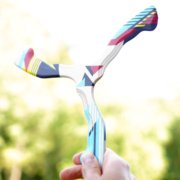 Ranku-darbo-bumerangas-laikomas-rankoje Vyro ranka laiko spalvotą medinį bumerangą „Uraganas“ žaliame gamtos fone.